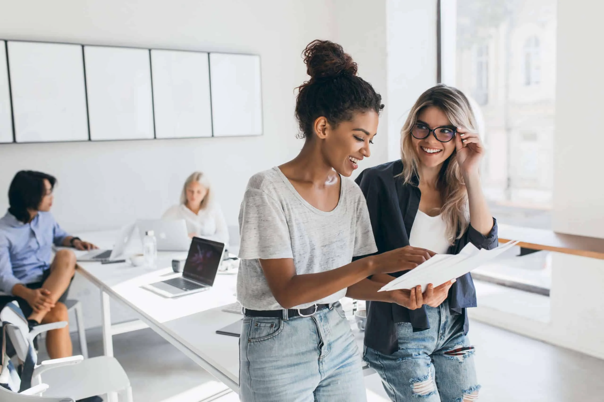 Gland blonde secretary in glasses looks at african female manager which holding report. Indoor portrait of stylish black woman in jeans spending time in office with asian and european colleagues..