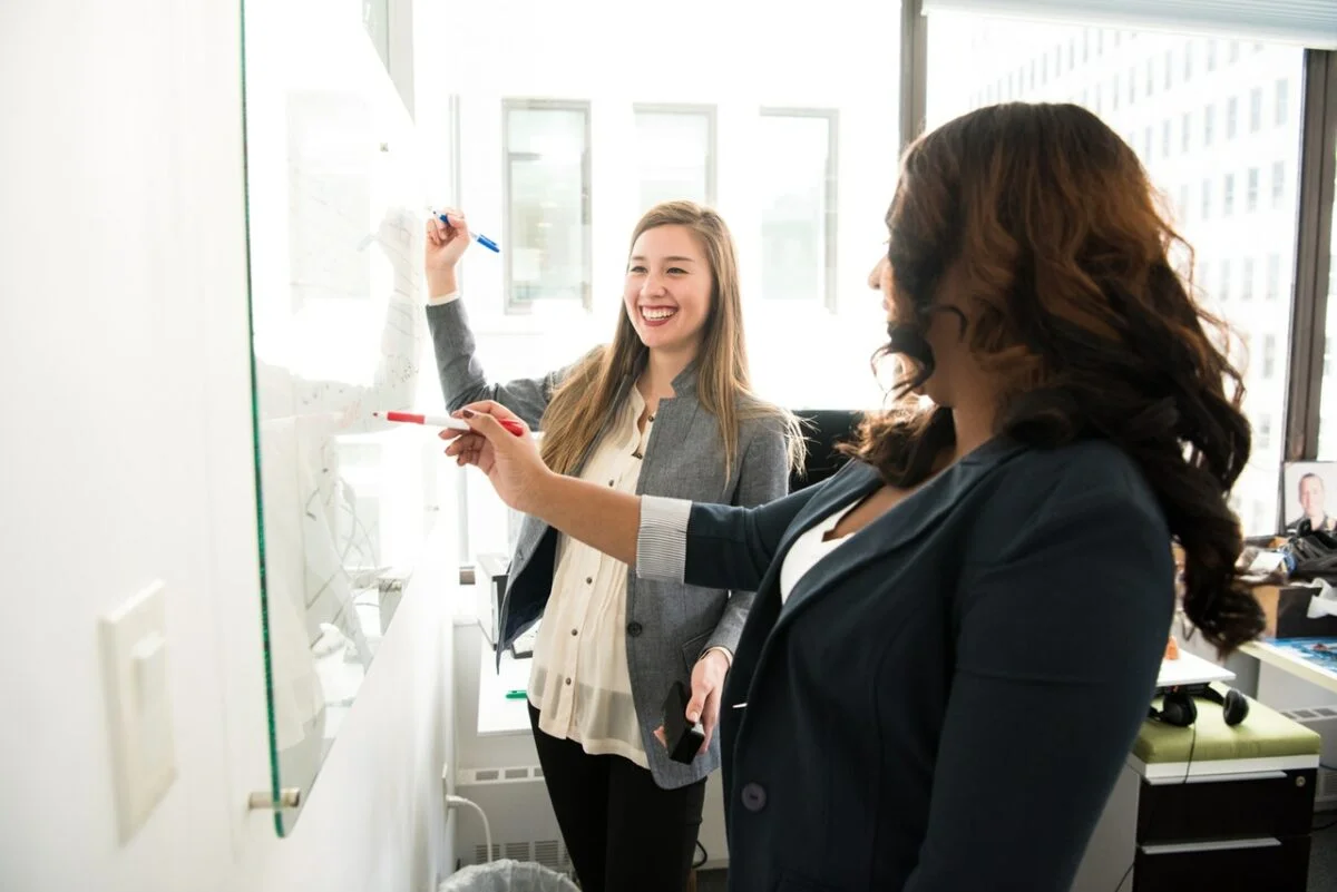 two woman working at a whiteboard with markers